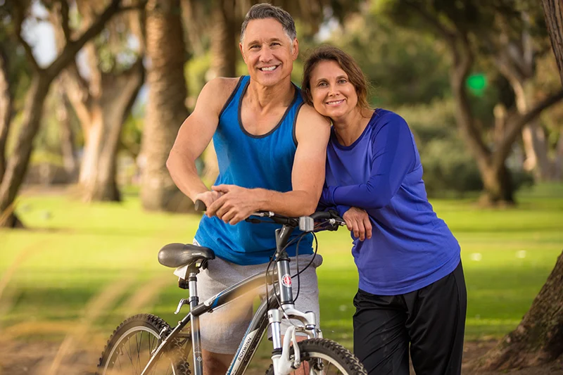 Smiling active middle-aged couple leaning on a bicycle together in a sunny park.