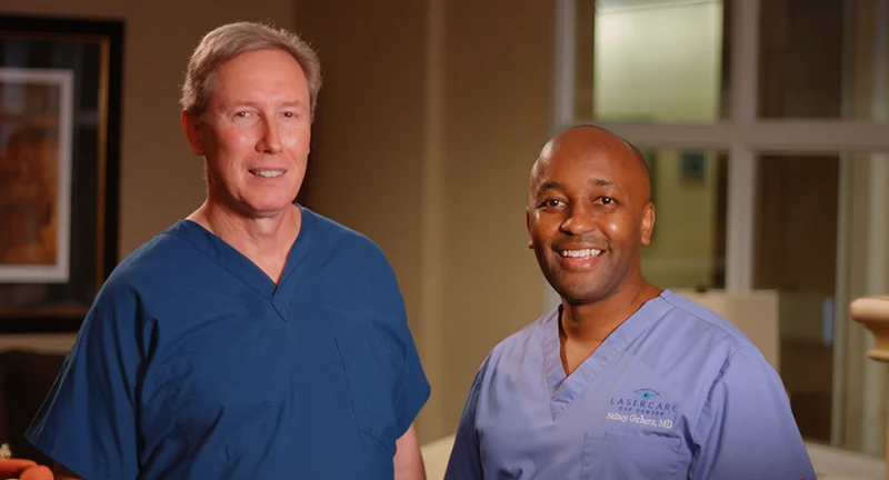 Dr. William Boothe and Dr. Sidney Gicheru smiling in blue surgical scrubs inside LaserCare Eye Center.