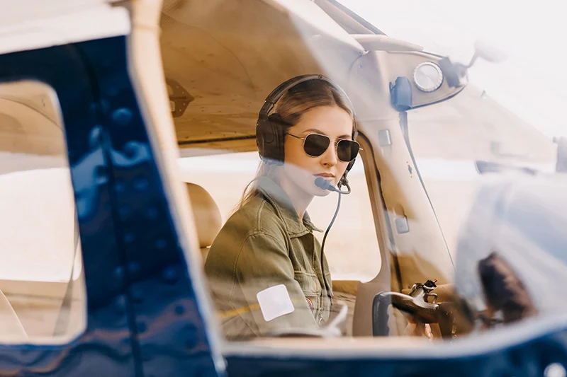 Female pilot wearing aviator sunglasses and a headset sitting inside an airplane cockpit.