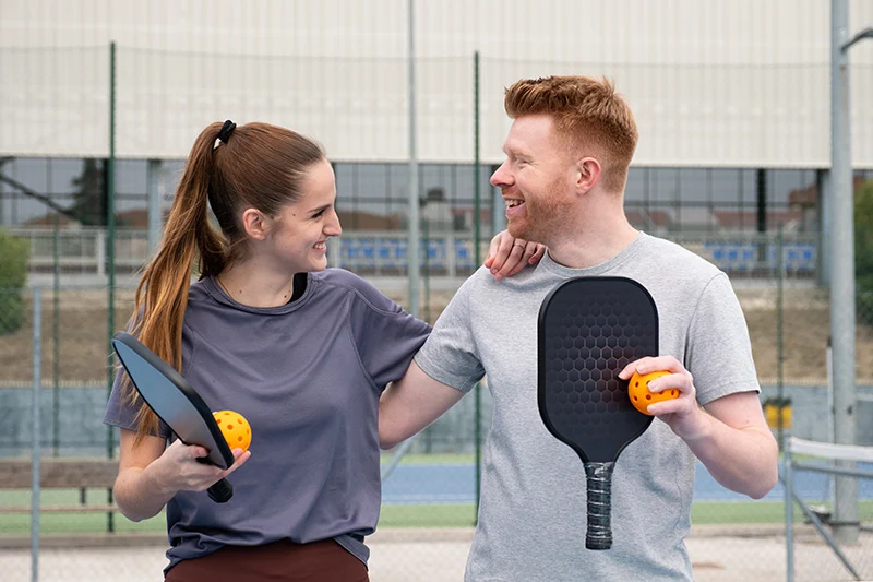 Smiling man and woman holding pickleball paddles and yellow pickleballs on an outdoor court.